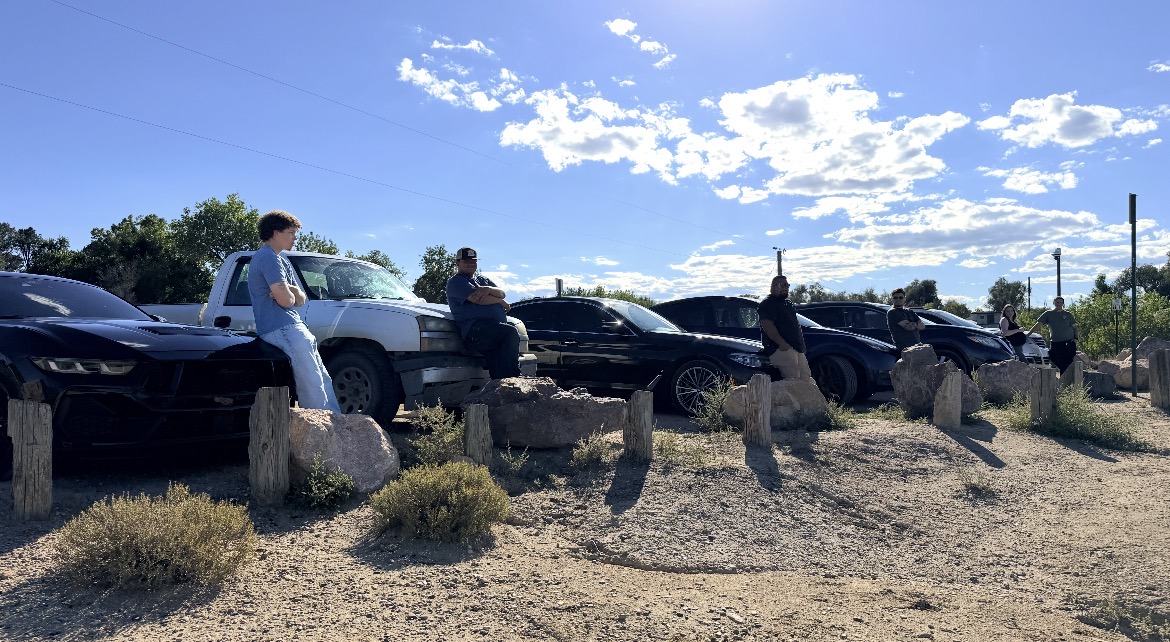 A row of members' cars parked on a dirt overlook under a blue sky, with people standing nearby